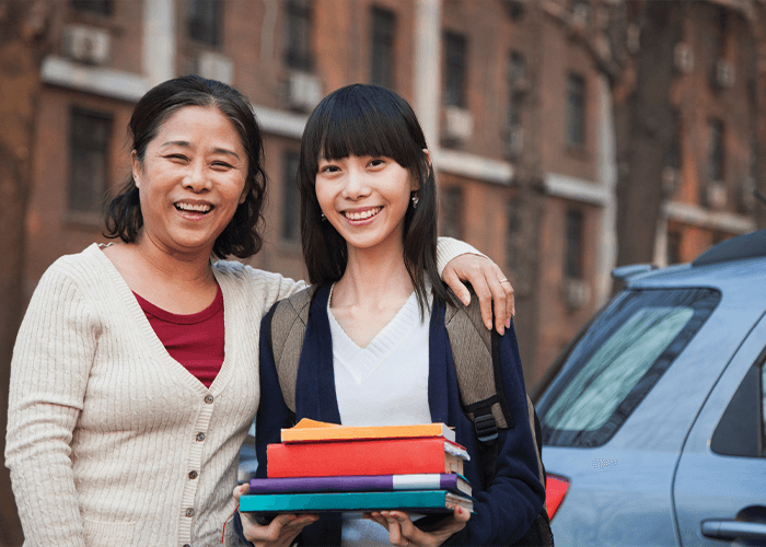 Asian mother and daughter smiling in front of a college dorm with moving boxes – stress-free student storage and moving made easy.